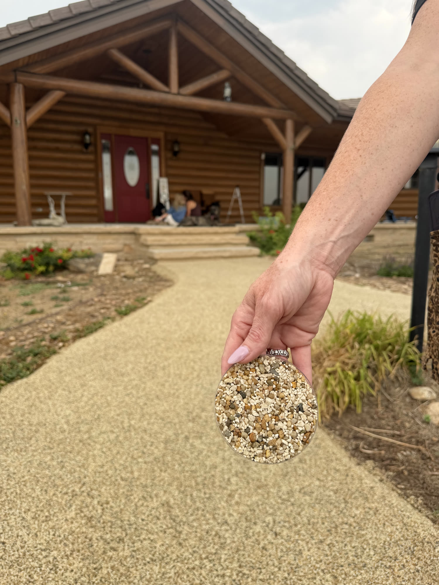 Hand holding a Vuba Stone sample in front of a finished tan walkway leading to a log home